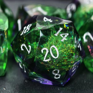 May include: A twenty-sided die featuring a translucent emerald green and deep purple resin body with internal iridescent foil flakes. The die has sharp geometric facets and engraved numbers painted in a bright silver color, with the number 20 visible.