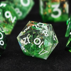 May include: A twenty-sided die made of translucent green resin with white numbers. The interior contains suspended green moss-like textures and small gold-colored metallic flakes. The die features sharp edges and clear, legible white numerals.