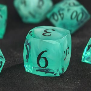 May include: A translucent teal six-sided die with black ink numbers. The die features a swirling internal pattern and sharp edges. The number nine is prominently displayed on the front face, with the number three visible on the top face.
