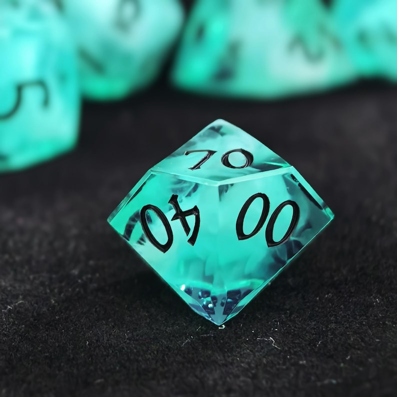 May include: A translucent, teal-colored ten-sided die featuring black engraved numbers. The die displays the numbers 70, 40, and 00 on its visible faces. The material has a clear, glass-like appearance with a smooth, multi-faceted surface.