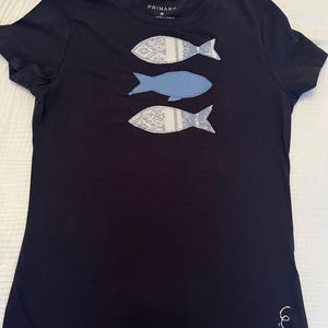 Könnte beinhalten: Marineblaues T-Shirt mit drei Fischapplikationen. Zwei Fische sind weiß mit einem Muster und einem hellblauen Streifen. Der mittlere Fisch ist einfarbig hellblau. Der Markenname "PRIMARK" ist am Ausschnitt sichtbar.