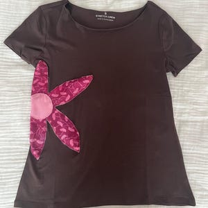 May include: A brown short-sleeved t-shirt featuring a large flower appliqué on the left side. The flower has a pink centre and petals made of patterned magenta fabric. The neckline displays the text "STRETCH CREW".