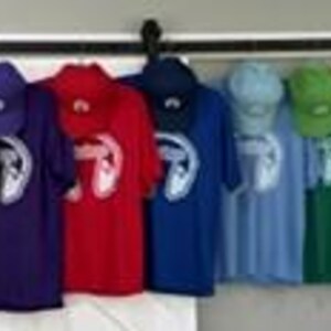 May include: A collection of short-sleeved t-shirts and matching baseball caps in a rainbow of colors including black, purple, red, blue, light blue, green, yellow, orange, and pink, each featuring a white graphic of headphones on the chest.
