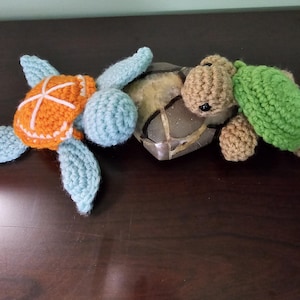 May include: Two handmade crochet sea turtles. One turtle has an orange shell with white accents, blue flippers and head. The other turtle has a tan head and flippers, and a green shell. The turtles are resting on a dark brown surface.