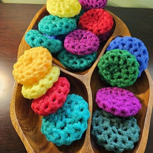 May include: A wooden serving tray filled with colorful, crocheted dish scrubbers. The scrubbers are in various colors, including yellow, red, teal, purple, green, and blue. The scrubbers are round and have a textured appearance, suggesting they are handmade.