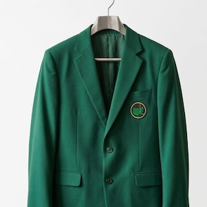 May include: A dark green blazer with a two-button closure, notched lapels, and two front pockets. A circular patch with a golf course design is on the chest. The jacket is hanging on a silver hanger.