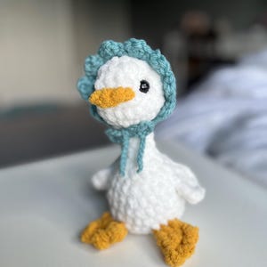 May include: A white crocheted duck plush toy featuring a textured body, orange feet, and an orange beak. The duck wears a blue crocheted bonnet with a scalloped edge and two dangling ties under the chin. It has a single visible black plastic eye.