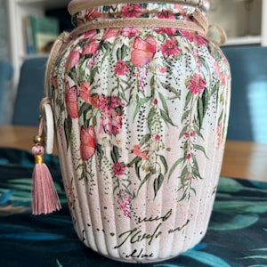 May include: A decorative jar with a lid, decorated with a floral pattern of pink flowers and green leaves. The jar is wrapped with twine and has a pink tassel. The text on the jar reads "Kindness Travels".