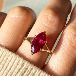 Puede incluir: Un anillo de oro con una gran piedra preciosa de color rojo intenso en talla marquesa, montada en un engaste de garras. La banda tiene un diseño dividido que se conecta a la base de la piedra, mostrando un acabado metálico pulido.