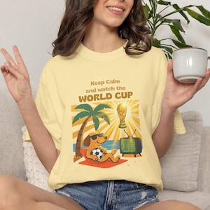 May include: Light yellow t-shirt with the text "Keep Calm and watch the WORLD CUP." The graphic features a dog wearing sunglasses, a soccer ball, a palm tree, a TV, and the World Cup trophy. The shirt is being worn by a person.