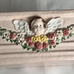Könnte beinhalten: Ein rechteckiger, hellrosa Pflanzkübel mit einem zentralen Relief eines Cherubgesichts mit weißen Flügeln, umgeben von einer Girlande aus modellierten rosa und gelben Rosen, weißen Blüten und grünen Blättern sowie zwei dekorativen Quasten.