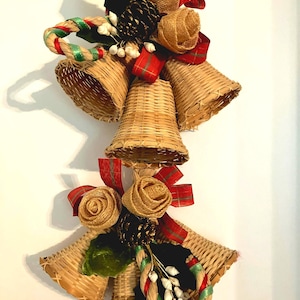 May include: A decorative hanging ornament featuring four woven wicker bells arranged in a cluster. The bells are accented with tan burlap rosettes, dark brown pinecones, green leaves, white berries, and red and green plaid fabric ribbons.