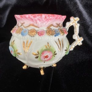 May include: A white porcelain footed cup featuring a scalloped pink rim, a handle with gold accents, and a decorative band of small blue, pink, and gold flowers. The body of the cup is adorned with additional floral motifs and gold leaf detailing.