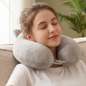 May include: A grey travel neck pillow with ear-shaped details is around a person's neck. The pillow is made of soft material and is designed for comfort. The person is wearing a white t-shirt and is sitting on a beige sofa.
