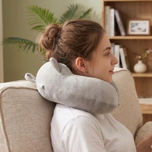 May include: A grey travel pillow in the shape of a cartoon animal, with small ears and a loop. The pillow is around the neck of a person sitting on a sofa.