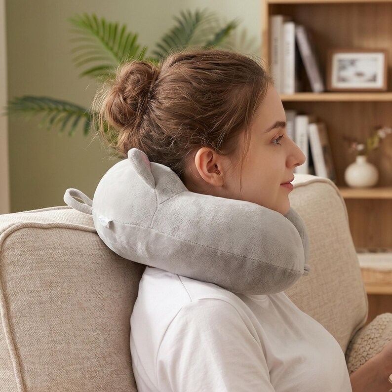 May include: A grey travel pillow in the shape of a cartoon animal, with small ears and a loop. The pillow is around the neck of a person sitting on a sofa.