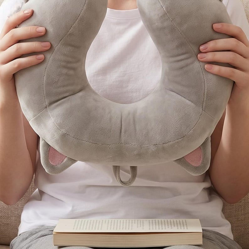 May include: A grey travel pillow in the shape of a cat's head, featuring pink inner ears and a small loop. The pillow is held in front of a person wearing a white t-shirt. A book is in front of the person.