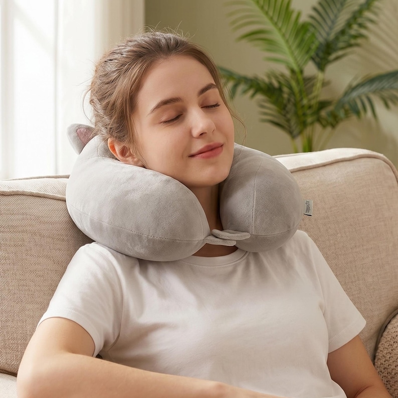 May include: A grey travel neck pillow with ear-shaped details is around a person's neck. The pillow is made of soft material and is designed for comfort. The person is wearing a white t-shirt and is sitting on a beige sofa.