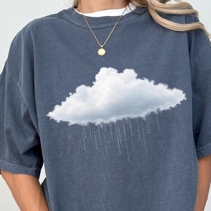 Może przedstawiać: Ciemnoniebieski t-shirt z krótkim rękawem z nadrukiem białej, puszystej chmury z pionowymi liniami przedstawiającymi padający deszcz na środku klatki piersiowej. Koszulka ma okrągły dekolt, luźny krój oraz złoty naszyjnik z łańcuszka.