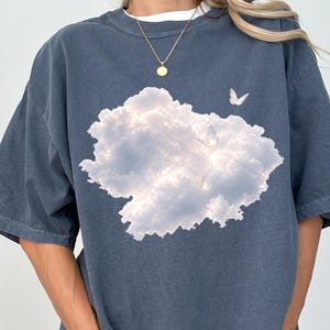 May include: A dark slate blue short-sleeve t-shirt featuring a large, central graphic print of a fluffy white cloud formation against a soft sky. Several small, delicate white butterflies are scattered across the cloud graphic, adding detail.