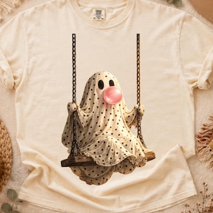 Puede incluir: Una camiseta de manga corta color crema con el gráfico de un fantasma cubierto por una sábana blanca con lunares negros, sentado en un columpio de madera suspendido por cadenas, mientras sopla una burbuja redonda de color rosa brillante.