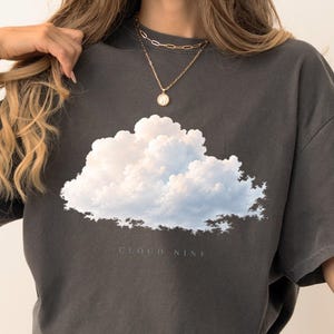 May include: A charcoal grey short-sleeve t-shirt featuring a large, realistic white and light blue cloud graphic centred on the chest. Below the cloud, the text CLOUD NINE is printed in a subtle, small, and clean sans-serif font.