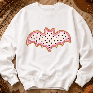 May include: A white long-sleeve sweatshirt featuring a central graphic of a bat. The bat has a cream-colored body with black polka dots, outlined by a pink border and a shimmering gold glitter edge, creating a festive and decorative appearance.