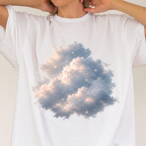 May include: A white short-sleeved t-shirt featuring a central graphic print of a large, fluffy cumulus cloud. The cloud is rendered in shades of soft blue, grey, and white, with small, bright, golden-yellow star-like sparkles scattered throughout.