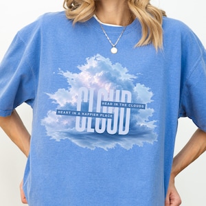May include: A blue short-sleeved t-shirt featuring a graphic of white clouds and the text CLOUD in large letters. Smaller text reads HEAD IN THE CLOUDS and HEART IN A HAPPIER PLACE. The shirt has a crew neckline and a relaxed, loose-fitting silhouette.