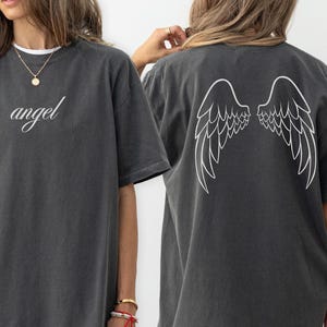 Op de afbeelding: Donkergrijs t-shirt met het woord "angel" in cursiefschrift op de voorkant. De achterkant van het shirt heeft een wit vleugelontwerp. Het shirt is een casual, oversized stijl.