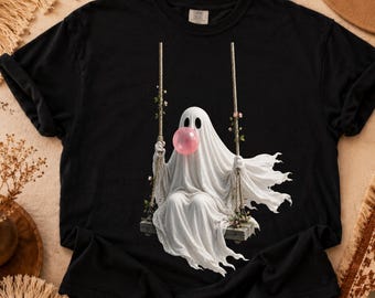 May include: A black short-sleeved t-shirt featuring a graphic print of a white ghost sitting on a wooden swing. The ghost has large black eyes and is blowing a round pink bubblegum bubble. The swing ropes are decorated with small floral vines.