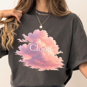 Puede incluir: Una camiseta de manga corta gris carbón con un gráfico central grande de nubes de atardecer en tonos rosa y naranja. La palabra Cloud está impresa en letras mayúsculas blancas sin remates en el centro de la ilustración de nubes frontal.
