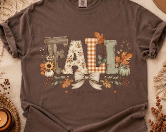 Puede incluir: Una camiseta de manga corta de color marrón oscuro con la palabra FALL en letras decorativas grandes. Las letras tienen patrones de vetas de madera, flores, cuadros vichy naranjas y verde sólido. Un lazo color crema, calabazas y hojas decoran el diseño.