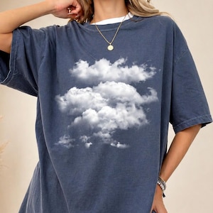 Könnte beinhalten: Dunkelblaues T-Shirt mit weißer Wolkengrafik. Das Shirt hat kurze Ärmel und eine lockere Passform. Das Model trägt ein weißes Unterhemd und eine goldene Halskette. Das Shirt ist aus einem weichen, bequemen Material.