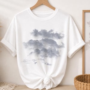 May include: White t-shirt featuring a cloudy sky design. The graphic shows a cluster of gray clouds against a white background. The shirt is hanging on a wooden hanger, with the sleeves rolled up.