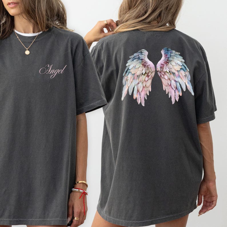 May include: Dark gray t-shirt with the word "Angel" in cursive on the front. The back of the shirt features a colorful watercolor design of angel wings in shades of pink, blue, and white. The shirt has a relaxed fit.