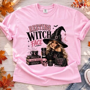 May include: A light pink t-shirt featuring a graphic of a person wearing a black witch hat, holding a coffee cup labeled Witch Fuel. The design includes a stack of books, a skull, a candle, a handbag, and the text Resting Witch Face.
