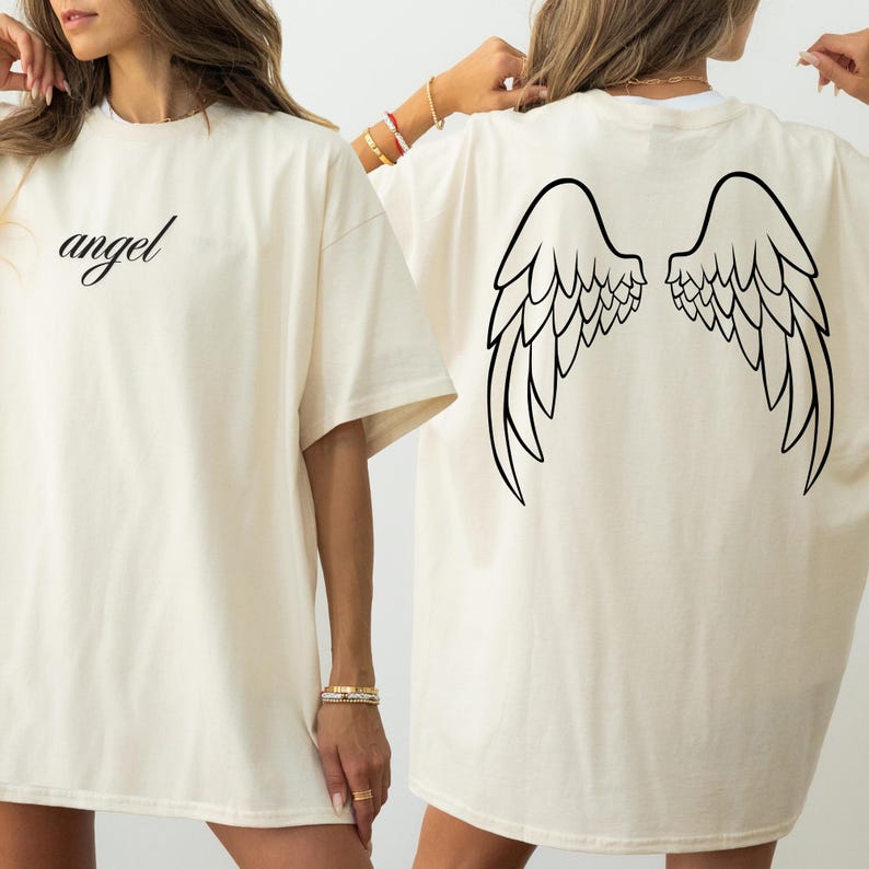 Op de afbeelding: Cr&egrave;mekleurig oversized T-shirt met het woord "angel" in een scriptlettertype op de voorkant. De achterkant van de shirt heeft een zwart vleugelontwerp. Het shirt heeft korte mouwen en een relaxte pasvorm.