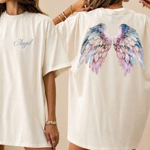 May include: Cream-colored t-shirt with the word "Angel" in script on the front. The back of the shirt features a watercolor design of angel wings in shades of blue, pink, and purple. The shirt has short sleeves and a relaxed fit.