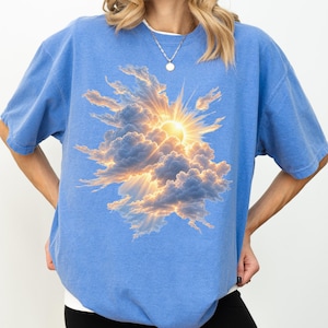 May include: A blue short-sleeved t-shirt featuring a graphic print of a bright sun shining through a cluster of fluffy clouds. The sun emits golden rays that extend outwards, illuminating the edges of the clouds against the blue fabric background.