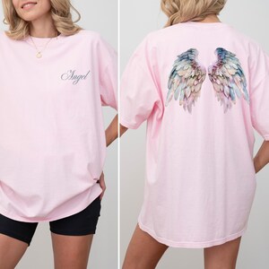 May include: A light pink t-shirt with the word "Angel" in cursive on the front. The back of the shirt features a watercolor design of angel wings in shades of blue, green, and pink. The shirt has short sleeves.