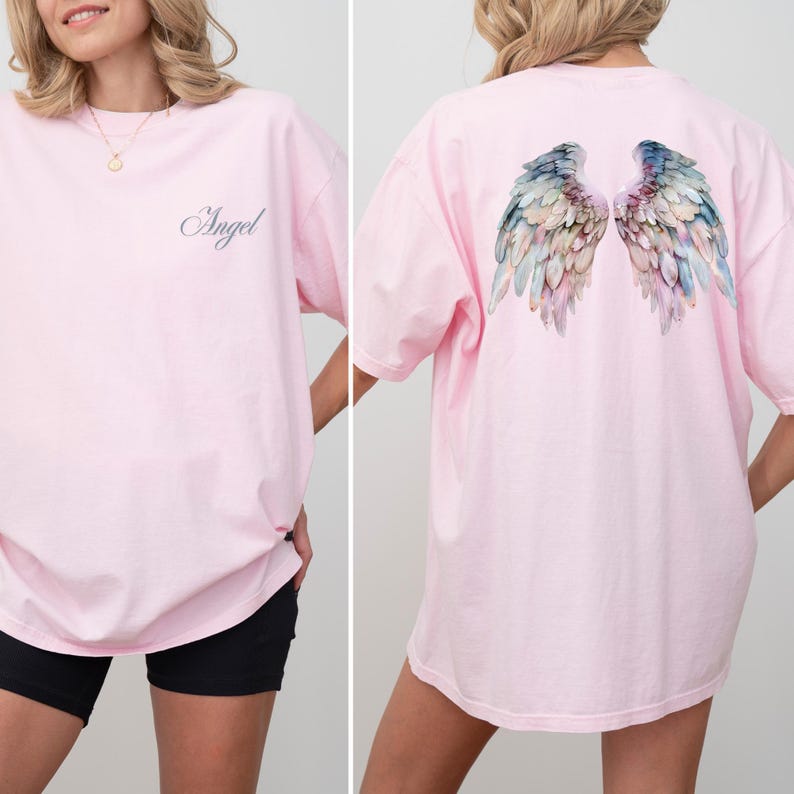 May include: A light pink t-shirt with the word "Angel" in cursive on the front. The back of the shirt features a watercolor design of angel wings in shades of blue, green, and pink. The shirt has short sleeves.
