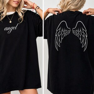 Op de afbeelding: Zwart oversized T-shirt met het woord "angel" in cursiefschrift op de voorkant. De achterkant van het shirt heeft een witte afbeelding van engelen vleugels. Het shirt heeft een ronde hals en korte mouwen.