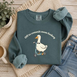 May include: Teal sweatshirt featuring the text "Off to commit mom foolery" in white script, with a cartoon duck on a light blue scooter. The sweatshirt has a relaxed fit and long sleeves. The sleeves are rolled up.
