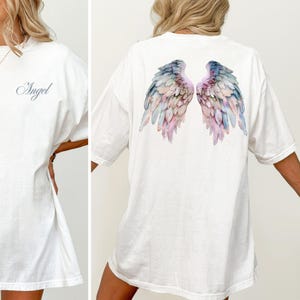 May include: White t-shirt with the word "Angel" in cursive on the front. The back of the shirt features a watercolor design of angel wings in shades of blue, pink, and purple. The shirt is a casual, oversized fit.