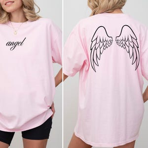 Op de afbeelding: Een lichtroze t-shirt met het woord "angel" in cursiefschrift op de voorkant. De achterkant van de shirt heeft een zwarte omtrek van engelen vleugels. Het shirt is een casual, oversized stijl.