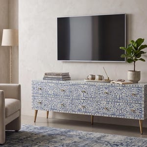 May include: A white and blue floral patterned cabinet with gold legs and hardware. The cabinet is in a living room setting with a TV, plant, and books. The room has a neutral color palette.
