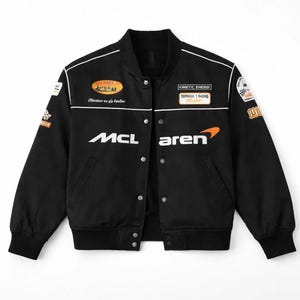 Könnte beinhalten: Eine schwarze College-Jacke mit weißer Paspelierung, Druckknopfleiste und gerippten Bündchen. Auf der Brust befindet sich der Schriftzug McLaren in Weiß und Orange. Verschiedene Rennsport-Aufnäher und Schriftzüge sind auf Brust und Ärmeln zu sehen.