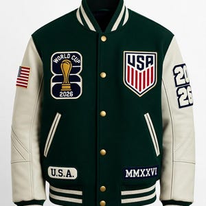 Op de afbeelding: Een donkergroen varsity-jack met crèmekleurige mouwen, voorzien van een World Cup 2026 trofee-patch, een USA-schildembleem, een kleine Amerikaanse vlag en de tekst U.S.A. en MMXXVI. Het jack heeft gouden drukknopen en gestreepte geribbelde boorden.