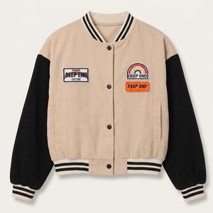 Op de afbeelding: Een beige corduroy varsity-jack met zwarte mouwen, een gestreepte kraag en boorden. De voorzijde is voorzien van patches met de tekst "HAWAII DEEP END CATTEE" en "DEEP END" met een regenboogafbeelding. Het jack heeft een drukknoopsluiting.